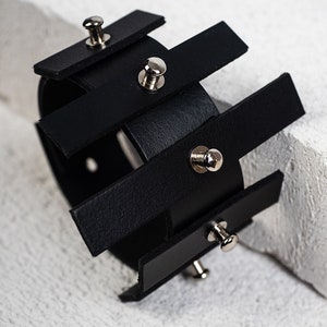 Puede incluir: Pulsera de cuero negro con tachuelas plateadas. La pulsera est&aacute; hecha de varias tiras de cuero que se superponen y se sujetan con tachuelas plateadas.