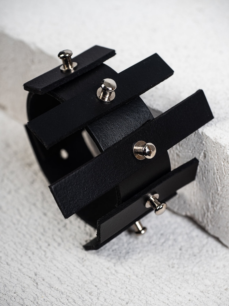 Puede incluir: Pulsera de cuero negro con tachuelas plateadas. La pulsera est&aacute; hecha de varias tiras de cuero que se superponen y se sujetan con tachuelas plateadas.