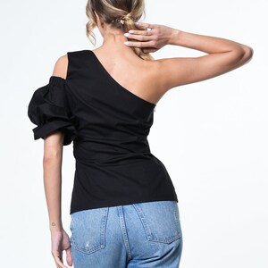 May include: A black one-shoulder top with a puffed sleeve and a fitted bodice. The top is paired with blue denim jeans.