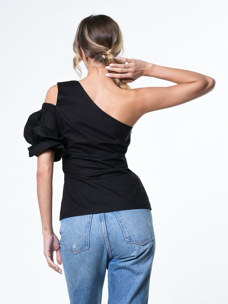May include: A black one-shoulder top with a puffed sleeve and a fitted bodice. The top is paired with blue denim jeans.