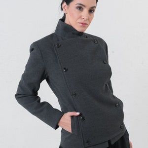 May include: A charcoal gray jacket with a high collar and asymmetrical button closure. The jacket features multiple dark buttons and a pocket. The model is wearing black pants. The jacket is a stylish piece of outerwear.