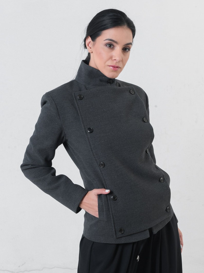 May include: A charcoal gray jacket with a high collar and asymmetrical button closure. The jacket features multiple dark buttons and a pocket. The model is wearing black pants. The jacket is a stylish piece of outerwear.