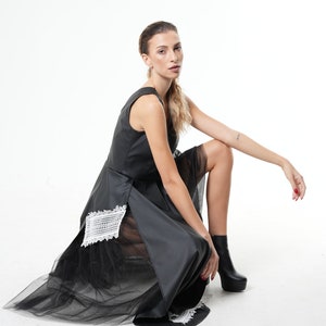 May include: A black sleeveless dress with a sheer tulle overlay and white lace detailing. The dress has a high neckline and a fitted bodice. The model is wearing black ankle boots.