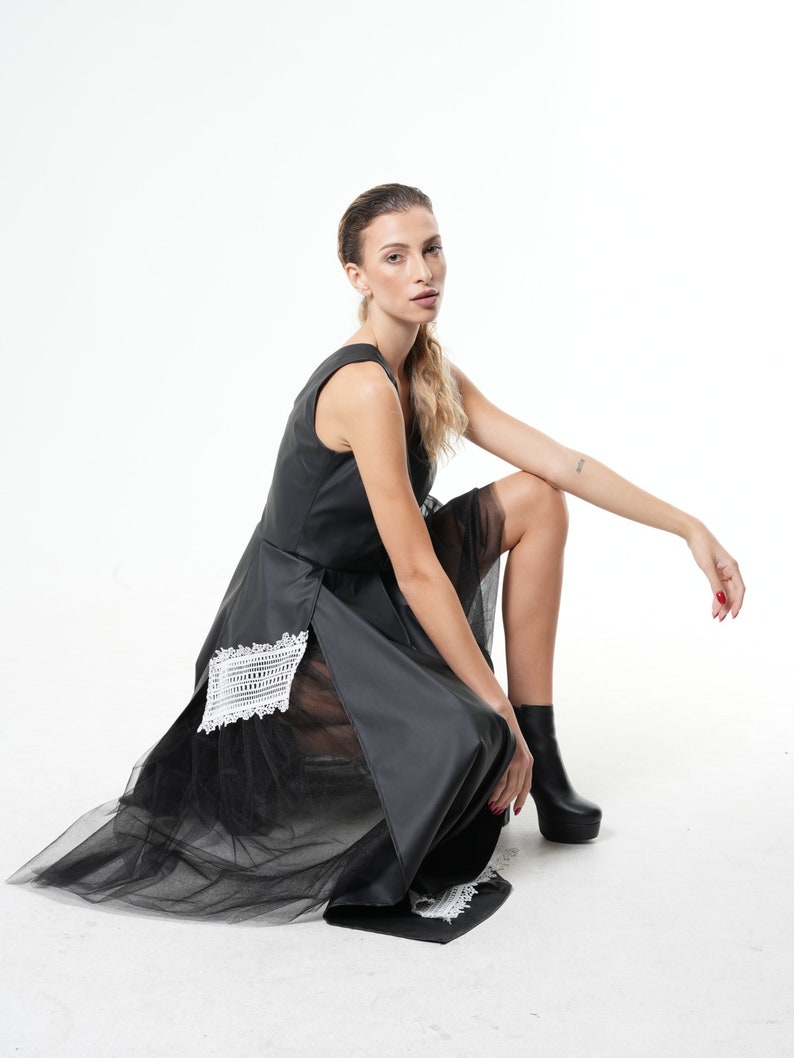 May include: A black sleeveless dress with a sheer tulle overlay and white lace detailing. The dress has a high neckline and a fitted bodice. The model is wearing black ankle boots.