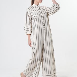 May include: A white and green striped jumpsuit with a button-up front and long sleeves. The jumpsuit has a wide leg and a relaxed fit. The model is wearing a brown cap.