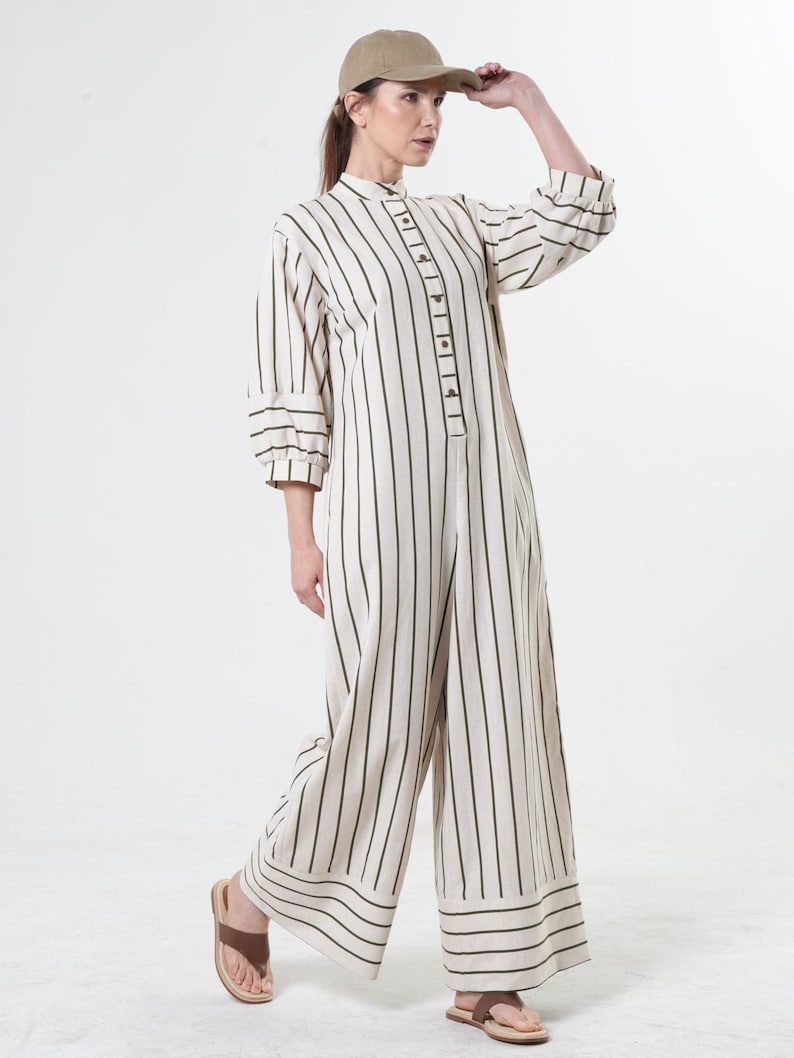 May include: A white and green striped jumpsuit with a button-up front and long sleeves. The jumpsuit has a wide leg and a relaxed fit. The model is wearing a brown cap.