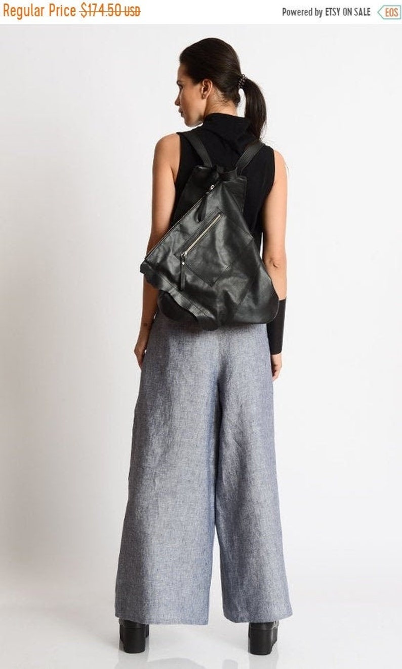 May include: A black leather backpack with a zippered pocket and a flap closure. The backpack is being worn by a person wearing a black tank top and wide-legged blue pants.