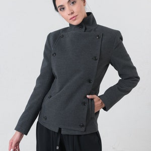 May include: A charcoal gray double-breasted jacket with a high collar and black buttons. The jacket has a tailored fit and a pocket on the front. The model is wearing black pants.