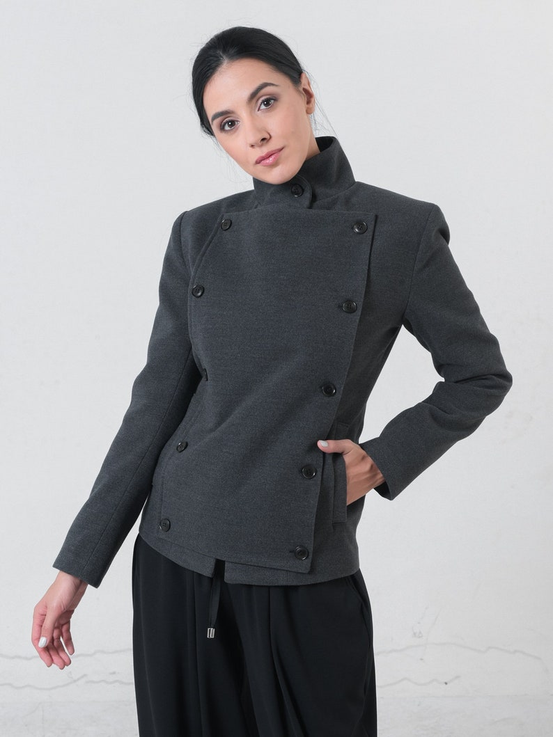 May include: A charcoal gray double-breasted jacket with a high collar and black buttons. The jacket has a tailored fit and a pocket on the front. The model is wearing black pants.