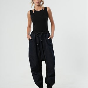 May include: A woman is wearing a black tank top and dark blue denim pants with a loose, wide-leg fit. The pants have a drawstring waistband and a unique design with a gathered section at the crotch.