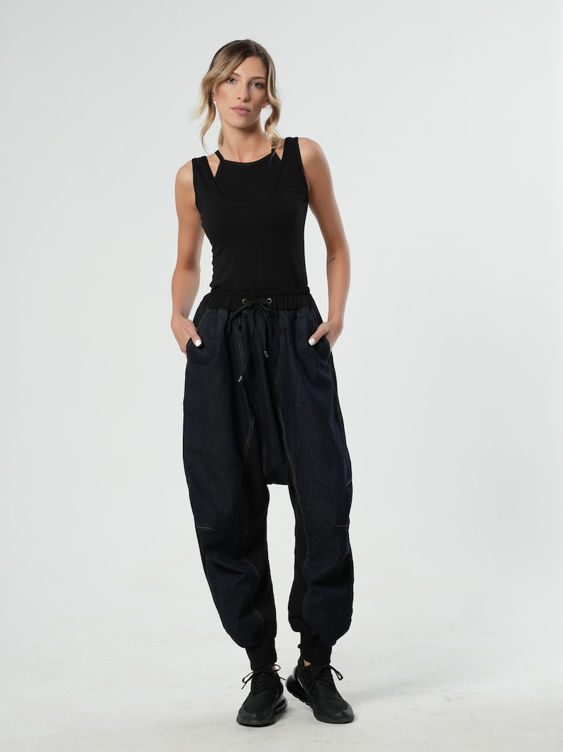 May include: A woman is wearing a black tank top and dark blue denim pants with a loose, wide-leg fit. The pants have a drawstring waistband and a unique design with a gathered section at the crotch.