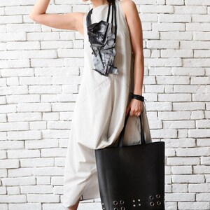 May include: A woman wearing a light gray sleeveless dress with a black geometric necklace and black sunglasses. She is holding a large black tote bag with silver grommets and wearing black fur-trimmed slippers.