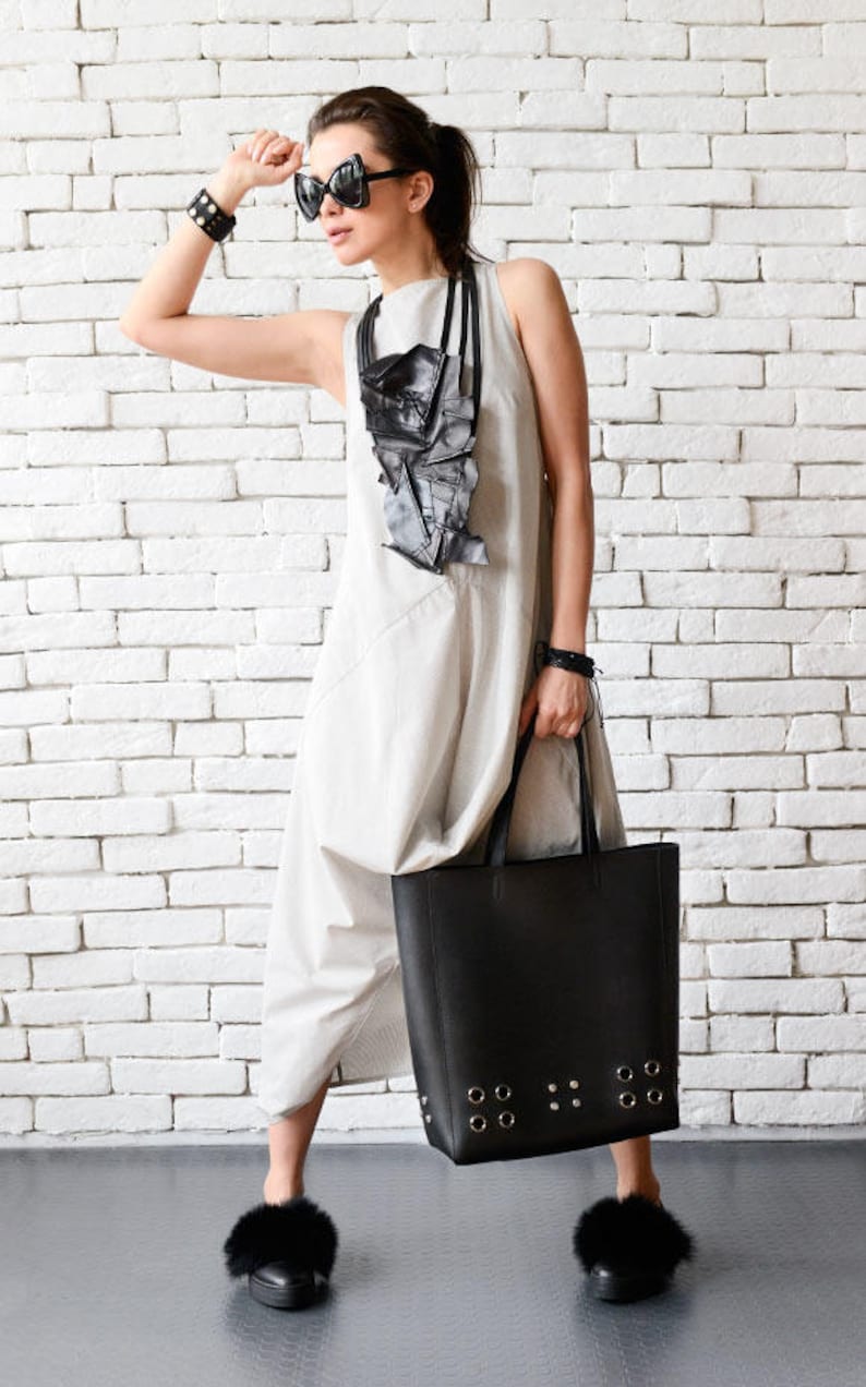 May include: A woman wearing a light gray sleeveless dress with a black geometric necklace and black sunglasses. She is holding a large black tote bag with silver grommets and wearing black fur-trimmed slippers.