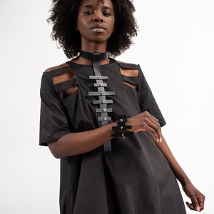 Puede incluir: Un vestido negro con recortes y un collar y pulsera de cuero. El vestido tiene un corte holgado y manga corta.