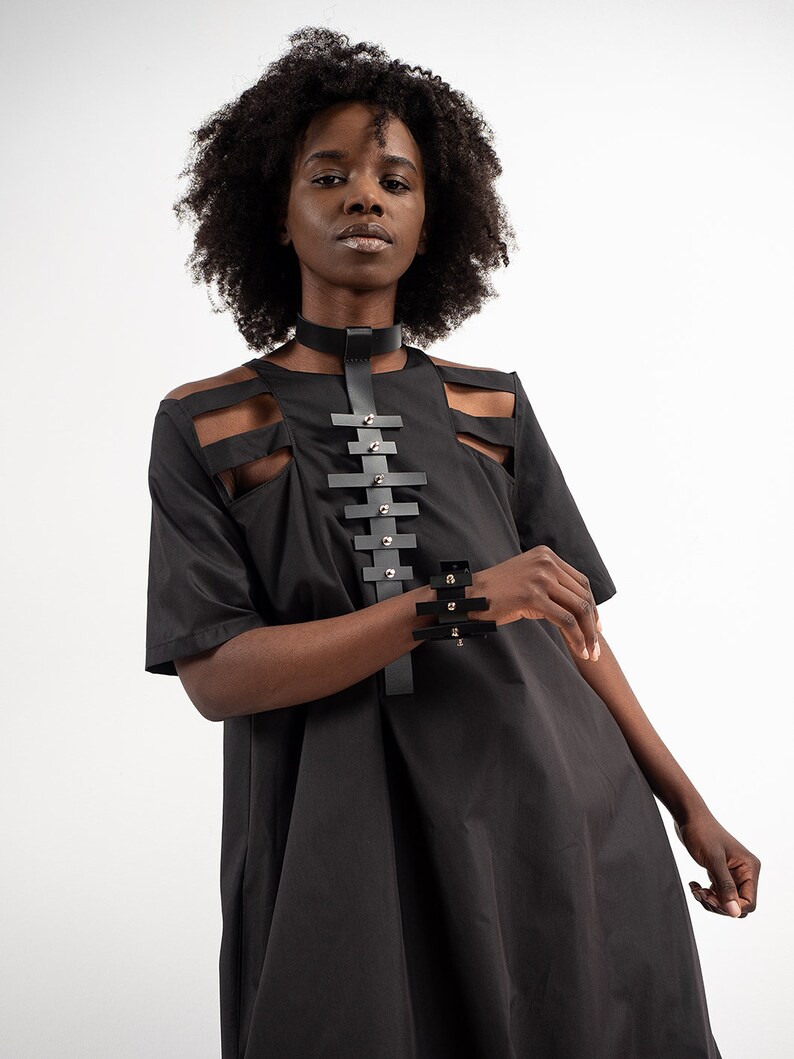 Puede incluir: Un vestido negro con recortes y un collar y pulsera de cuero. El vestido tiene un corte holgado y manga corta.