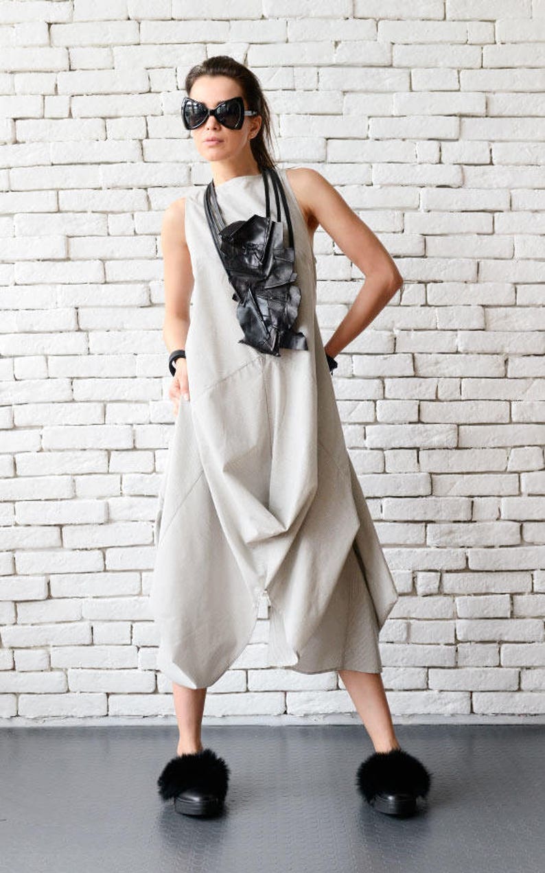 May include: A woman wearing a gray jumpsuit with a black leather necklace and black sunglasses. She is standing in front of a white brick wall and wearing black fur-trimmed shoes.