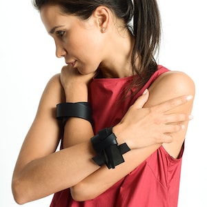 May include: A woman wearing a red sleeveless top with two black leather bracelets on her wrists. The bracelet on her left wrist is a simple band, while the bracelet on her right wrist is a knotted design.