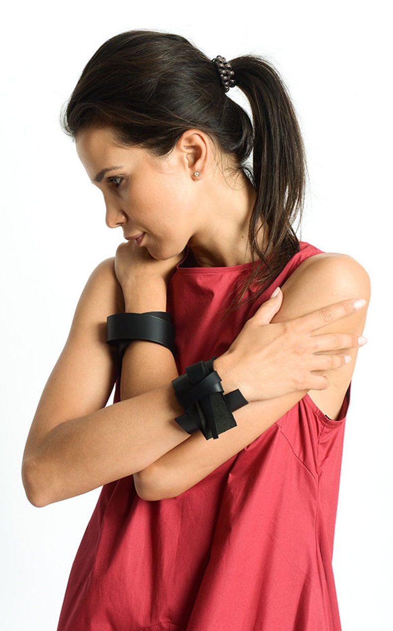 May include: A woman wearing a red sleeveless top with two black leather bracelets on her wrists. The bracelet on her left wrist is a simple band, while the bracelet on her right wrist is a knotted design.