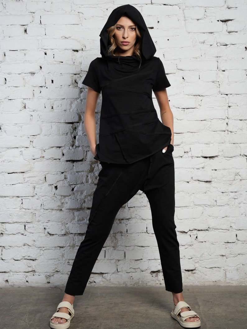 May include: A black hooded top with a layered design and a matching pair of black pants. The model is wearing the outfit with white sandals.
