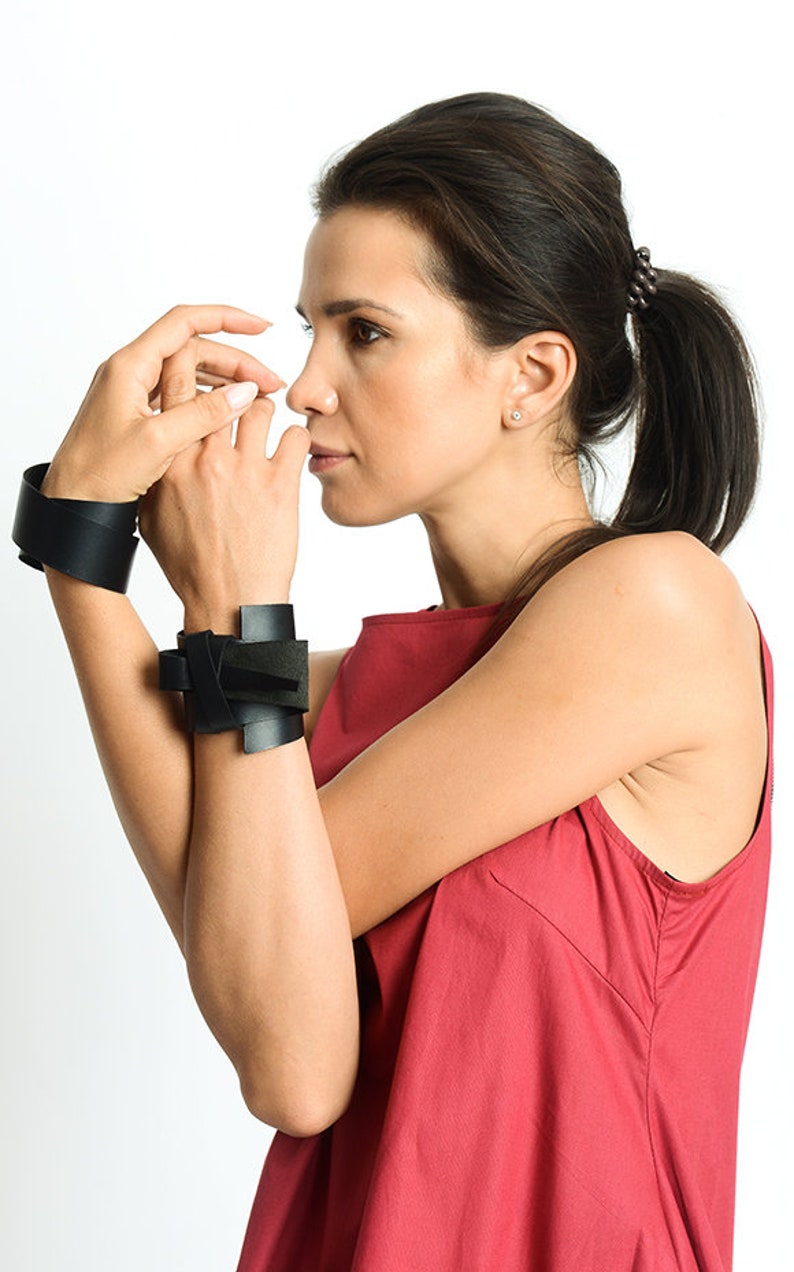 May include: Two black leather bracelets on a person's wrist. The bracelets are both wide and have a simple design.