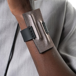 Può includere: Un bracciale a fascia larga caratterizzato da un pannello rettangolare color bronzo metallizzato con una cresta centrale rialzata. Il pannello è fissato da un cinturino nero liscio che avvolge il polso, unito con cuciture nere visibili.