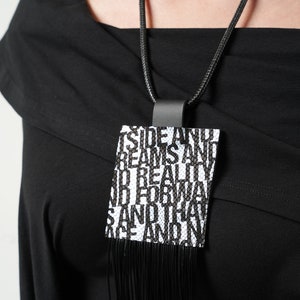Puede incluir: Collar negro con colgante rectangular con las palabras "Outside Dreams And Reality" en texto negro sobre fondo blanco. El colgante tiene flecos negros en la parte inferior y cuelga de un cord&oacute;n negro.