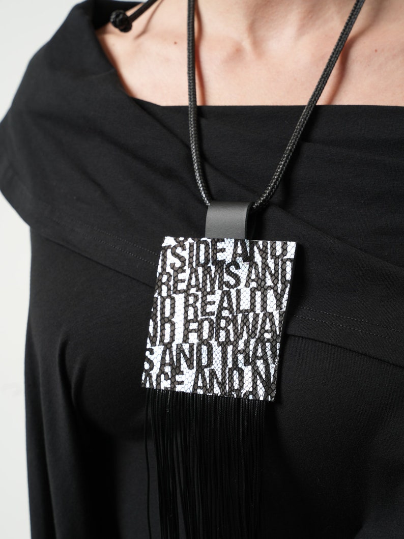 Puede incluir: Collar negro con colgante rectangular con las palabras "Outside Dreams And Reality" en texto negro sobre fondo blanco. El colgante tiene flecos negros en la parte inferior y cuelga de un cord&oacute;n negro.