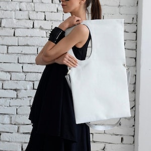 May include: A white leather tote bag with a large oval cutout. The bag has a long strap and is being held by a person wearing a black dress and sunglasses.
