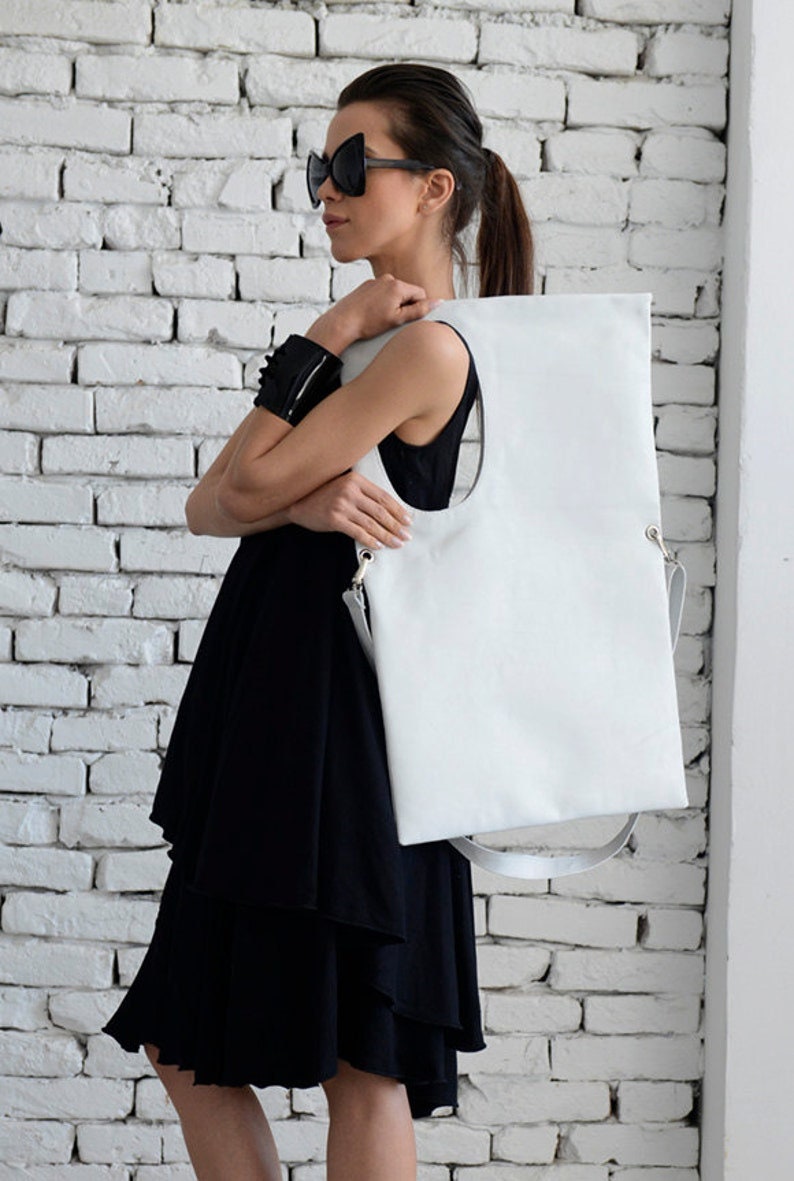 May include: A white leather tote bag with a large oval cutout. The bag has a long strap and is being held by a person wearing a black dress and sunglasses.