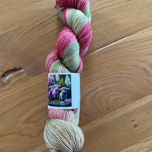 Op de afbeelding: Een streng garen in roze en groene tinten, gedraaid in een compacte vorm. Een label met cartoon schapen is om het garen gewikkeld. Het garen wordt gepresenteerd op een houten oppervlak.