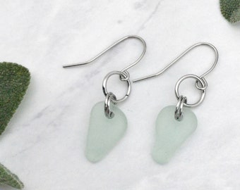 May include: A pair of silver earrings with light green sea glass teardrop charms. The earrings have a simple design and are perfect for everyday wear.