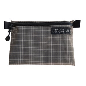 May include: A gray zippered pouch with a black zipper and a black strap. The pouch has a grid pattern and a black label that says "Riveted Oak Designs".