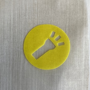 May include: A bright yellow, circular patch featuring a cut-out design of a flashlight. The flashlight graphic is positioned diagonally within the circle. The patch is made of a textured fabric.