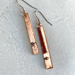 May include: A pair of copper earrings with a hammered texture and an arrow design. The earrings are attached to silver hooks.