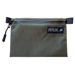 May include: A green zippered pouch with a black zipper and black straps. The pouch has a small black label with the text "Riveted Oak Designs" in white.