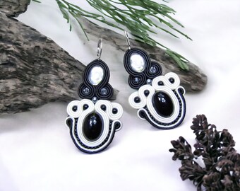 May include: Black and white dangling earrings with a black oval-shaped stone and a silver-toned bead in the center. The earrings are made of black and white string and have a silver hook.