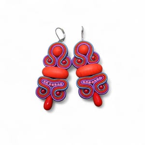 Op de afbeelding: Een paar handgemaakte, kleurrijke oorbellen. De oorbellen hebben een combinatie van rode, paarse, blauwe en roze soutache-koord. Elke oorbel heeft een grote rode kraal aan de bovenkant, een rode ovale kraal en een rode druppelkraal.