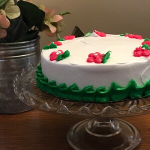 Puede incluir: Un pastel blanco con glaseado verde y rosa. El pastel está decorado con flores rosas y hojas verdes. El pastel está en un soporte para pastel de vidrio.