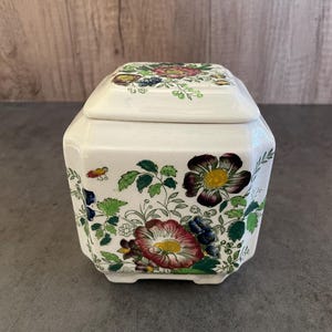 Könnte beinhalten: Eine quadratische weiße Keramikdose mit Deckel, verziert mit einem floralen Muster in Rot-, Gelb-, Grün-, Blau- und Violetttönen.