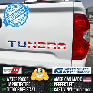 Peut inclure: Hayon de camion blanc avec le mot "TUNDRA" en couleurs patriotiques. Les lettres sont bleues avec des étoiles blanches et des rayures rouges. Le texte supplémentaire comprend "WATERPROOF", "UV PROTECTED", "OUTDOOR RESISTANT", "AMERICAN MADE", "PERFECT FIT", et "CAST VINYL (BUBBLE FREE)".