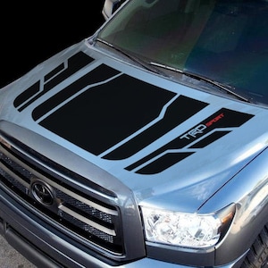 May include: A gray pickup truck with a black and gray "TRD Sport" hood decal. The truck has a black grille and headlights.