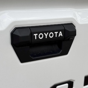 May include: A black plastic handle with a white "TOYOTA" logo on a white vehicle.