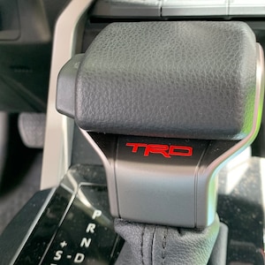 May include: Close-up of a black and silver car gear shift lever. The lever has a black leather top and the red letters "TRD" are visible on the silver base. The gear shift is in the "P" position.