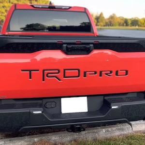 May include: Rear view of a bright orange Toyota Tundra TRD Pro pickup truck. The truck features black accents, including the tailgate, bumper, and trim. The text "TRD PRO" is prominently displayed on the tailgate.