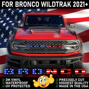 May include: Red Ford Bronco Wildtrak with a black grille and a red, white, and blue American flag design on the front. The text "FOR BRONCO WILDTRAK 2021+ " is at the top of the image. The text "BRONCO" is at the bottom of the image with a red, white, and blue American flag design. The text "3M VINYL", "WATERPROOF", and "UV PROTECTED" are listed below the text "BRONCO". The text "PRECISELY CUT", "HIGHEST QUALITY", and "MADE IN THE USA" are listed below the text "BRONCO" on the right side of the image.