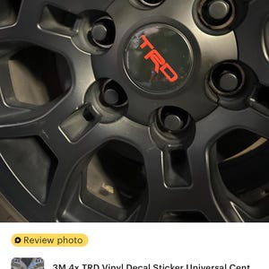 May include: A black car wheel with a red TRD logo decal. The wheel has five spokes and five lug nuts.
