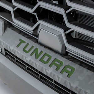 May include: A close-up of a grey truck grille with a black camera lens and the word "TUNDRA" in green letters.