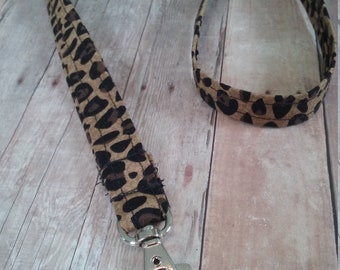 May include: A tan and brown leopard print lanyard with a silver-colored metal clasp. The lanyard is made of fabric and has a repeating pattern of dark brown spots on a tan background. The clasp is a swivel lobster claw style.