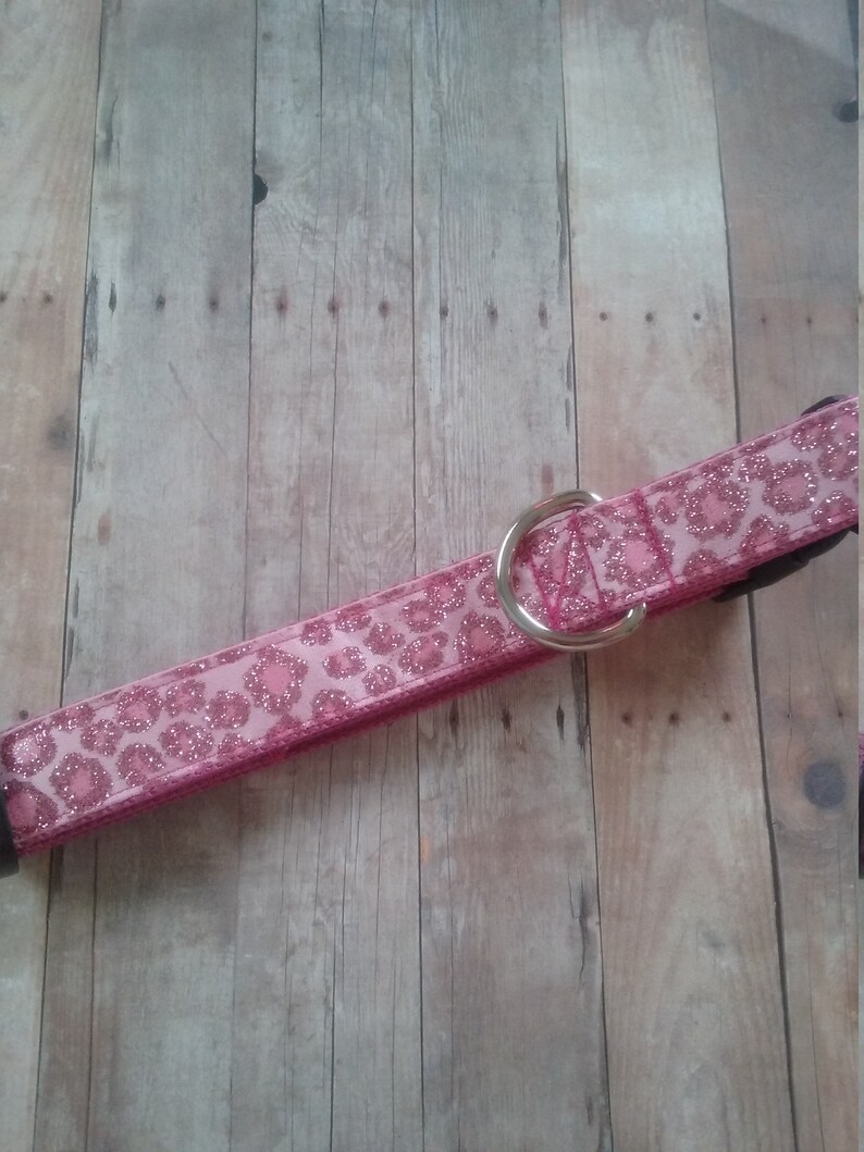 May include: Pink dog collar with a glittery leopard print design. The collar features a silver metal D-ring and a black buckle. The collar is made of a durable material and is designed for pets.