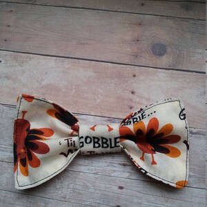 May include: A beige bow tie with a turkey print and the text "Til Gobble Til We Drop" in black.